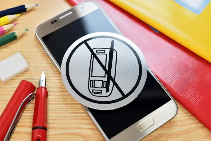 Handy mit Verbotsschild umgeben von Schulutensilien, Symbolfoto für Handyverbot in der Schule