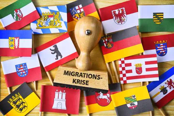 Stempel mit Aufschrift Migrationskrise inmitten Fahnen der deutschen Bundesländer, Symbolfoto überlastete Kommunen durch gestiegene Flüchtlingszahlen