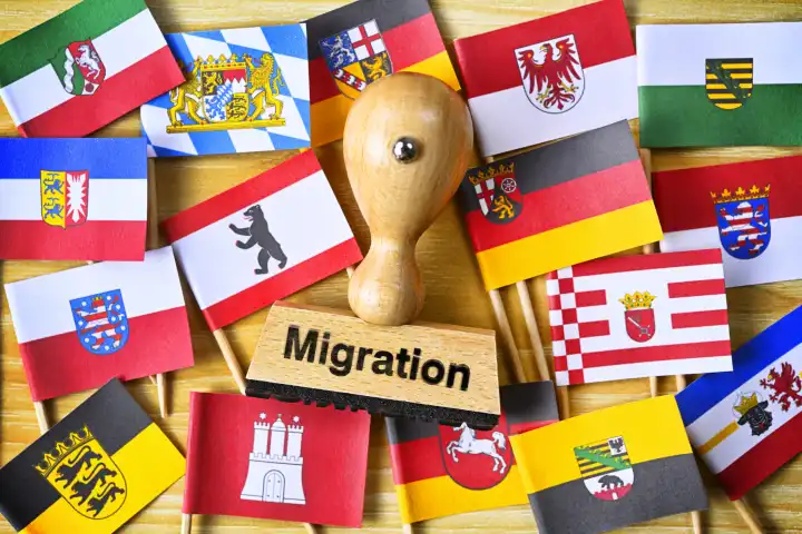 Stempel mit Aufschrift Migration inmitten Fahnen der deutschen Bundesländer, Symbolfoto überlastete Kommunen durch gestiegene Flüchtlingszahlen