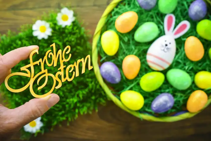 Hand hält den Schriftzug Frohe Ostern über bunten Ostereiern in einem Osternest