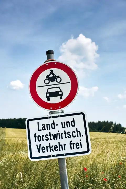 Traffic sign "Land- und Forstwirtsch. Traffic free" in wheat field against blue sky with white clouds and forest in background