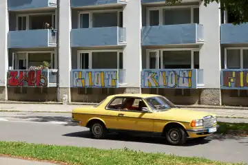 Gelber Mercedes BJ 1980er Jahre fährt vor 1970er Wohnblock mit Graffity