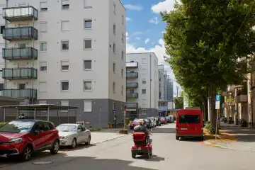 Roter fahrender Dreiradroller in der Krumpterstraße (MUC) zwischen Hochhäusern und zwei geparkten roten Fahrzeugen links und rechts von ihm