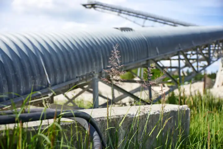 Conveyor belt of a gravel pit. Shuttering / metal wrapped on metal structure. Surrounded by green grasses. In the foreground a wall base on which two black rubber hoses lie.