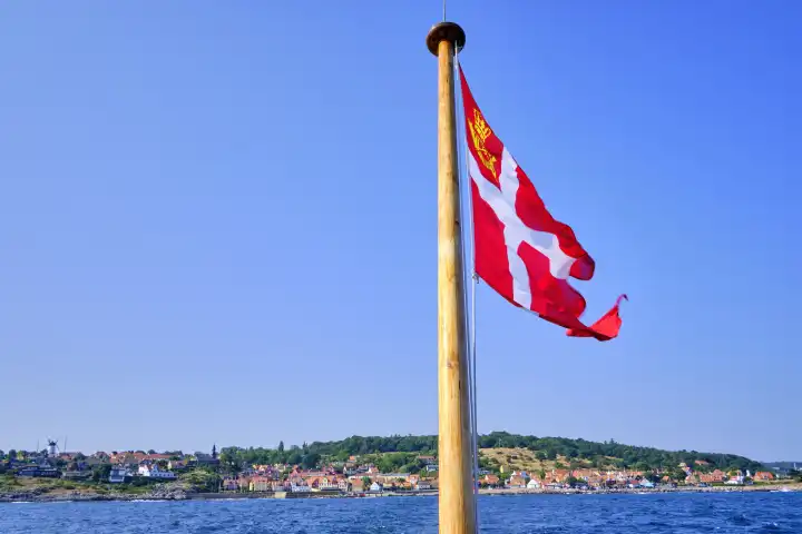 Wehende dänische Flagge am Heck eines Ausflugsschiffes und malerischer Blick auf die Küstenlandschaft von Gudhjem, Insel Bornholm, Dänemark.