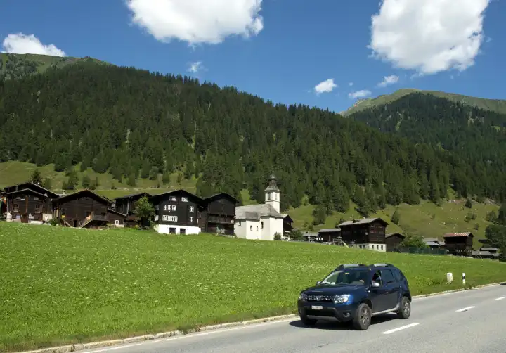 Bewegungsunscharfes Fahrzeug auf einer Landstrasse vor dem Ortsbild der Streusiedlung Gluringen, Goms, Wallis, Schweiz