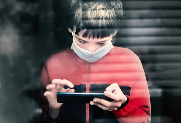 Teenage mit medizinischer Maske zu hause hinter einem Fenster benutzt sein Smart Phone