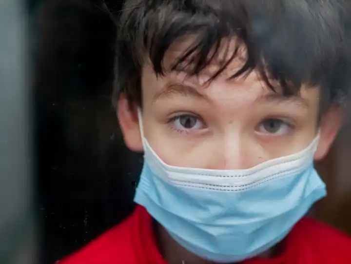 Teenage mit medizinischer Maske zu hause hinter einem Fenster