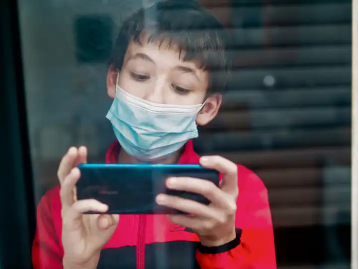 Teenage mit medizinischer Maske zu hause hinter einem Fenster benutzt sein Smart Phone