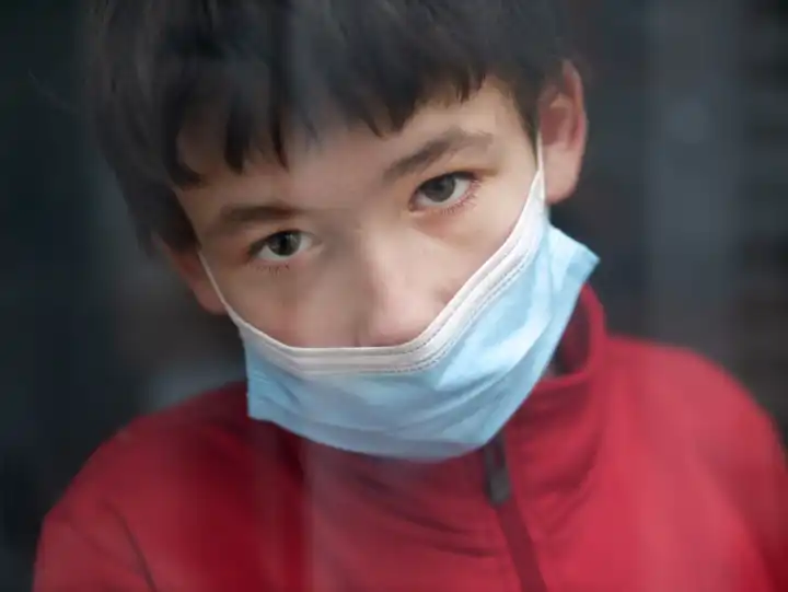 Teenage mit medizinischer Maske zu hause hinter einem Fenster