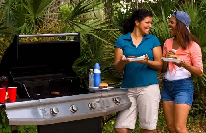 Two hispanic women enjoying picnic food outdoors at home with grill