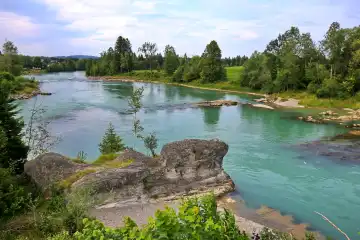 Das Flussufer des Lech in Lechbruck mit Blick auf eine Felsformation. Im Hintergrund befindet sich ein bewaldetes Gebiet mit Bäumen und einem blauen Himmel mit weißen Wolken. Das Bild hat eine friedliche und ruhige Stimmung. Lechbruck, Ostallgäu, Schwaben, Bayern, Deutschland.