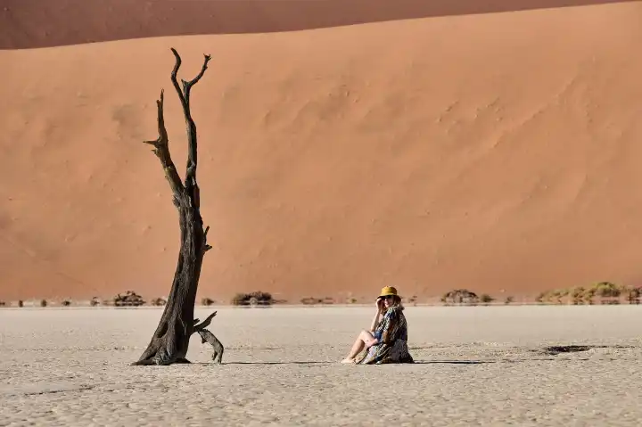 Young woman sits next to tree and in front of dune in Deadvlei