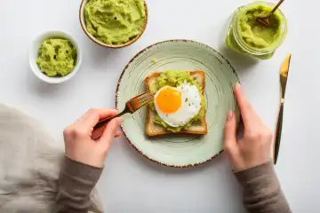 View of person eating fried egg and avocado puree on toast for breakfast, isolated on white, AI generated.