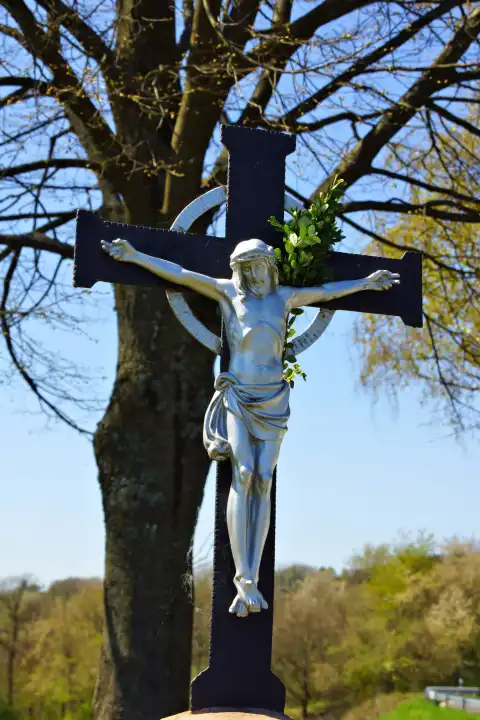 Kreuz mit Metallskulptur vor einem Baumstamm