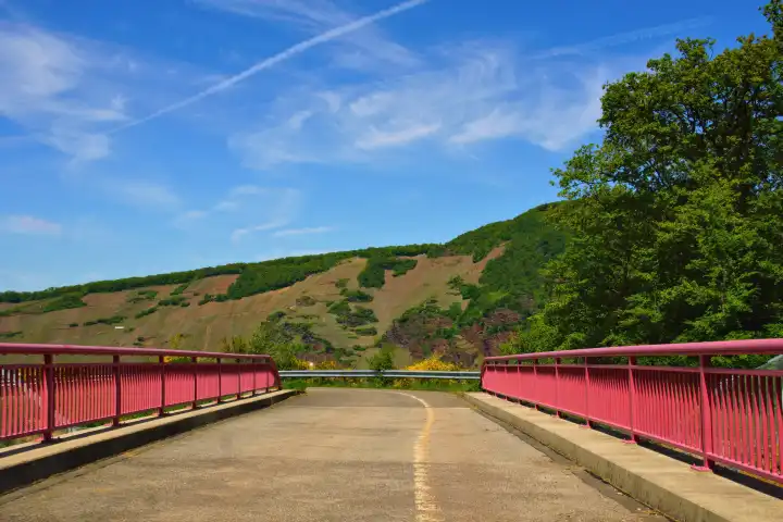 Brücke mit rotem Geländer vor den Felsen und den Weinbergen der Weinlage Ürziger Sonnenuhr