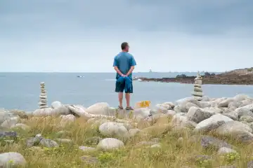 Mann steht zwischen zwei Steinmännchen und schaut aufs Meer