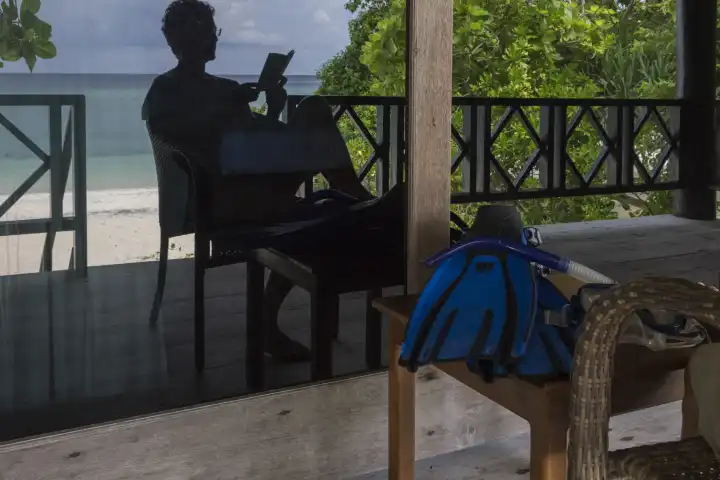 Spiegelbild einer lesenden Frau auf der Veranda, im Hintergrund Sandstrand und Meer