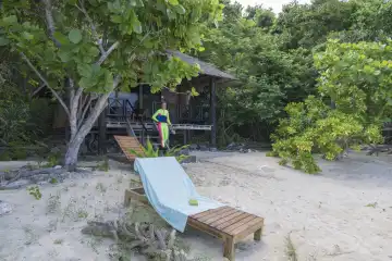 female snorkeler in front of wooden bungalow surrounded by lush shrubbery, in the foreground deck chair on sandy beach