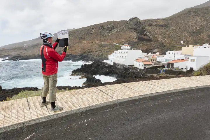 Woman taking pictures with tablet of churned sea and white houses of Tamaduste. El Hierro, Spain