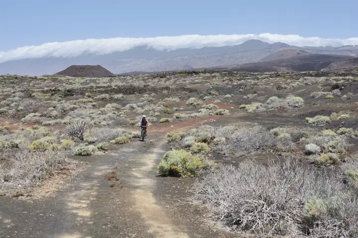 Mountainbikerin in karger, mit Pionierpflanzen bewachsener vulkanischer Landschaft. Blauer Himmel mit Wolkenband über Gebirge und Vulkankegel im Hintergrund. El Hierro, Kanarische Inseln, Spanien