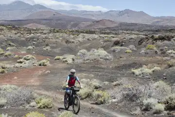 Mountainbikerin in karger, mit Pionierpflanzen bewachsener vulkanischer Landschaft. Blauer Himmel mit Wolkenband über Gebirge und Vulkankegel im Hintergrund. El Hierro, Kanarische Inseln, Spanien