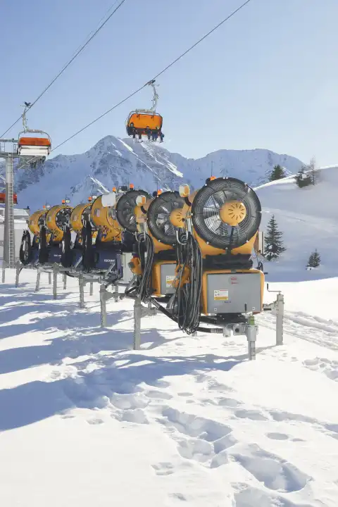 Mehrere Schneekanonen hintereinander aufgereiht mit Sesselbahn und Bergpanorama.