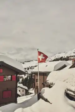 Flapping Swiss flag between snow-covered wooden chalets. Valais, Switzerland