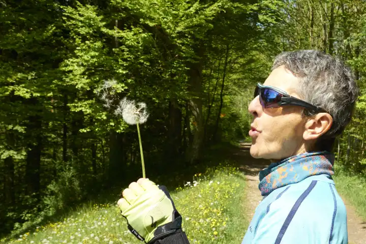 Sportliche Frau mit Sonnenbrille pustet die Samen einer Pusteblume weg. Zürcher Oberland, Schweiz