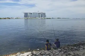 Two men fishing from the shore in a sea polluted with plastic waste. Blocks of flats are being built in the background. Makassar, Sulawesi, Indonesia