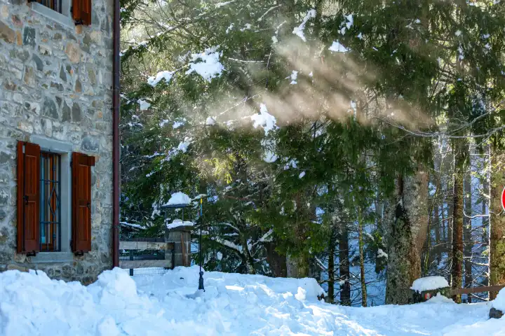 Winter- und schneereiche Landszene mit Steinhaus und Sonnenstrahlen, die durch die Bäume brechen.