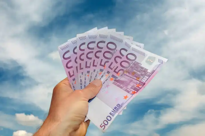 Hand hält einen Fächer mit 500 Euro Banknoten vor blauem Himmel