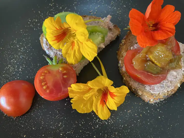 Snack consisting of bread halves with spreadable sausage, topped with red and green tomato slice, garnished with nasturtium flowers.