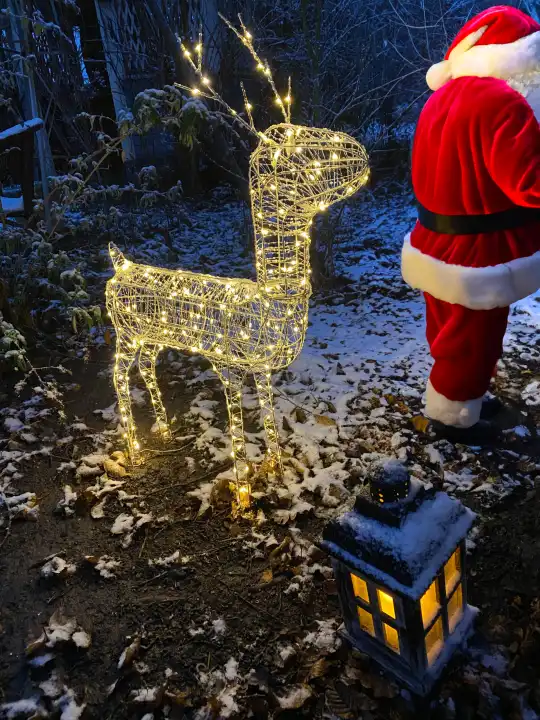 Own image generated with AI. Santa Claus with reindeer and lantern in the winter garden in the evening, Santa Claus is supplemented with AI
