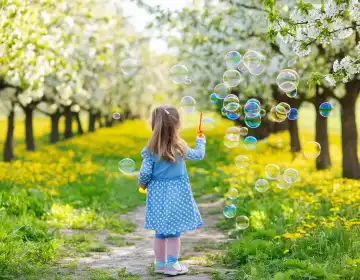 KI generiert. Kleines Mädchen, hellblau und rosa gekleidet, macht Seifenblasen auf einer Streuobstwiese zur Baumblütezeit