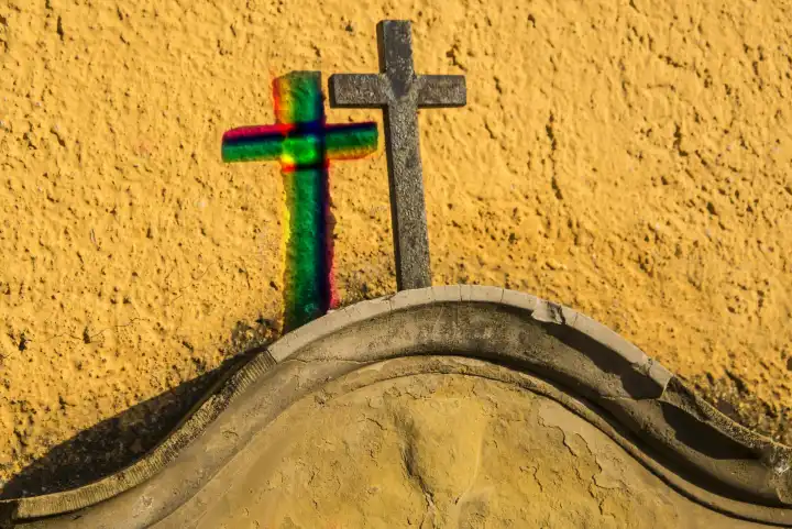 Kreuz auf einem Grabstein an einer gelben Wand mit Schatten in Regenbogenfarben