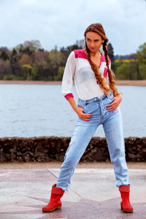 Young woman with long hair on lake shore in jeans and rubber boots in model pose