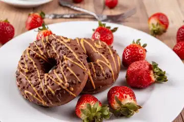 The picture shows two chocolate doughnuts on a white plate perched on a wooden surface. The doughnuts are garnished with fresh strawberries and arranged with cake forks beside them.