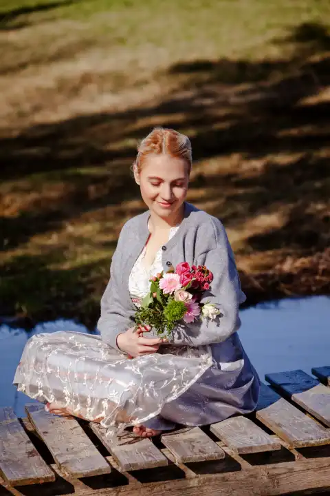 Eine junge Frau mit blonden Haaren sitzt lächelnd im Dirndl auf einem Holzsteg am Seeufer und hält Tulpen im Arm.