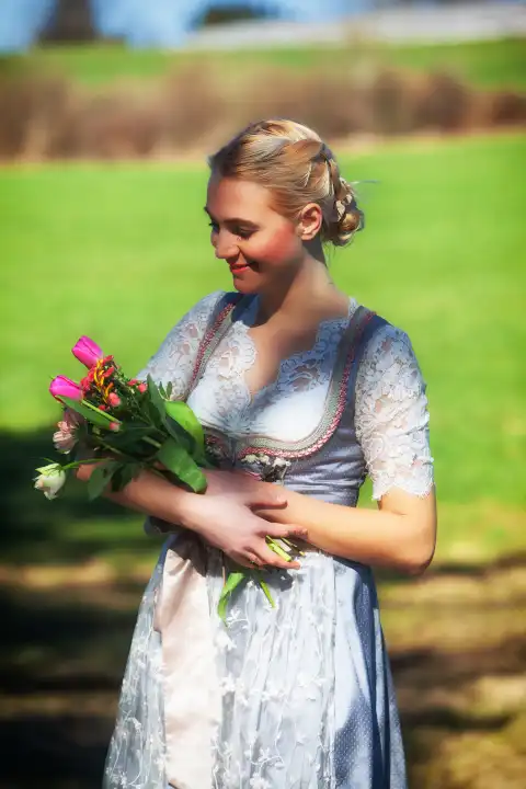 A young woman with blonde hair and a dirndl stands smiling in a flowering meadow, holding tulips in her arms.
