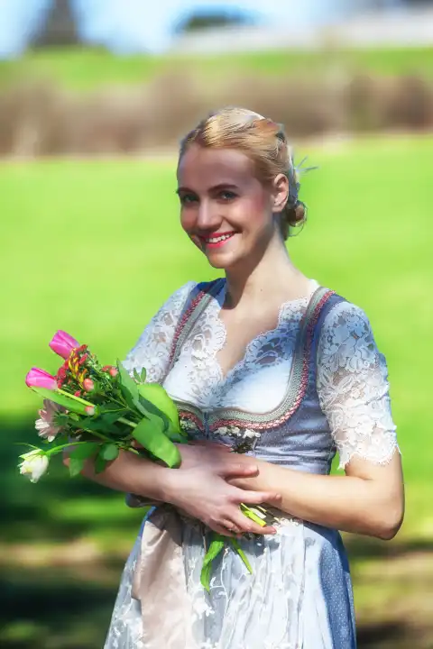 A smiling woman with blonde hair and a dirndl stands in a meadow, holding tulips and looking into the camera. 3/4 shot.