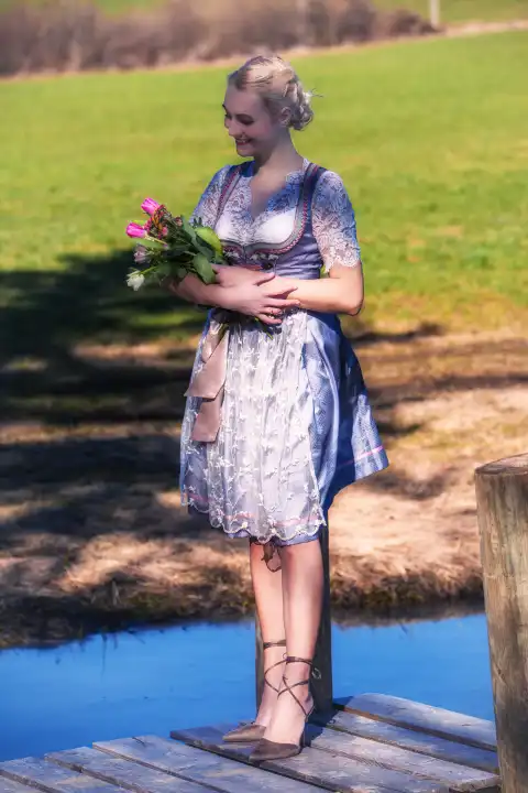 A smiling woman in a dirndl with blonde hair stands on a wooden footbridge over a lake, holding tulips in her arms.