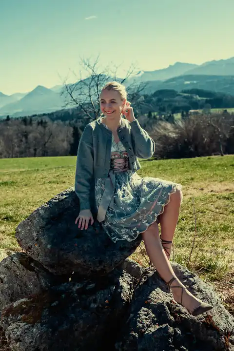 A woman with blonde hair and a dirndl sits on a stone in front of snow-covered mountains. A cardigan keeps her warm.