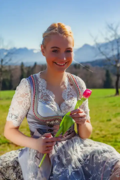A woman with blonde hair and a dirndl sits on a stone in front of snow-covered mountains in spring. She is holding a tulip in her hand.