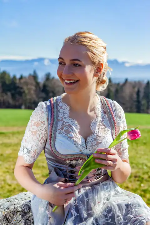 A woman with blonde hair and a dirndl sits on a stone in front of snow-covered mountains. She is holding a tulip and looking away to the side.