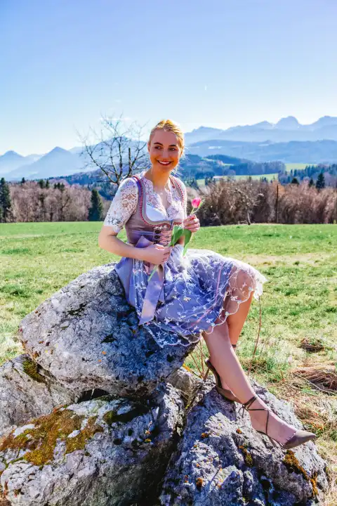 A woman with blonde hair and a dirndl sits smiling on a stone in front of snow-covered mountains, holding a tulip and beaming at the camera.