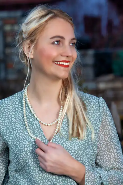 On a warm summer's day, a blonde woman in an airy summer dress stands in front of an idyllic backdrop. Her hair falls smoothly over her shoulders and her eyes shine in the sunlight. She wears an elegant pearl necklace around her neck, adding a touch of elegance to her outfit. With an enchanting smile, she radiates pure joie de vivre.