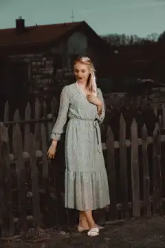 A young woman with blonde hair and a dreamy smile stands in front of an old wooden fence in an airy summer dress. The night air is warm and full of summer scents. A single ray of light falls on her from above and bathes her in a magical light. Her figure appears delicate and fragile in contrast to the rustic wood of the fence. The shadows of the night envelop her like a mysterious veil.