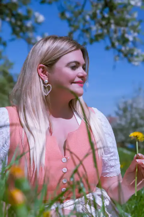 The picture shows a plus-size model in a vibrant orange dirndl sitting in a lush spring meadow. The sun scatters warm light across the terrain as the model smiles confidently and radiantly into the camera. The natural beauty of the scene is emphasized by the striking appearance of the model, whose blonde hair and voluptuous curves represent inspiring diversity.
