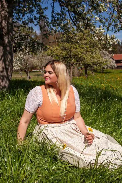 The picture shows a plus-size model sitting in a bright orange dirndl in a picturesque spring meadow. The sun bathes the scene in warm light, while the model smiles into the camera with a confident expression. The lush nature and the striking beauty of the model merge into a harmonious image full of life and color.